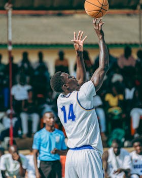 Athlete in blue and white jersey shooting a basketball during a lively outdoor game, crowd watching.