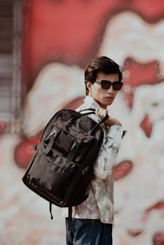 Stylish young man wearing sunglasses and backpack in front of graffiti wall.