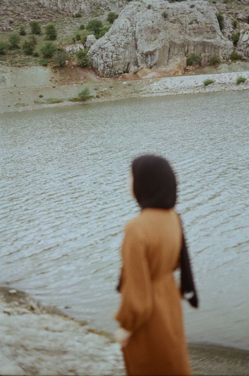 Free A woman in a brown dress standing by a tranquil lake with rocky hills. Stock Photo