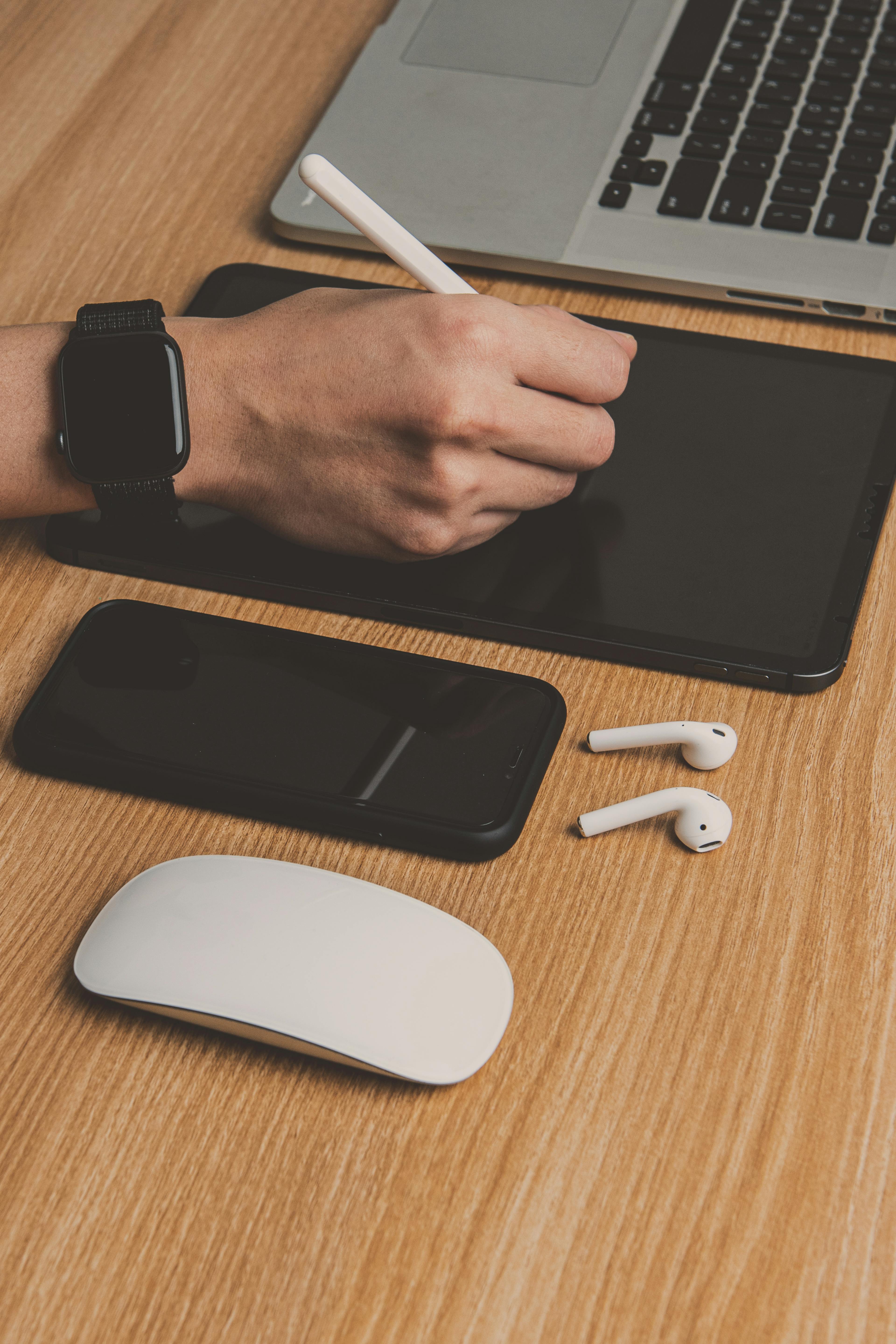 From above of crop unrecognizable person in smart watch using stylus and drawing tab while sitting near wireless mouse and earphones near laptop and smartphone on table