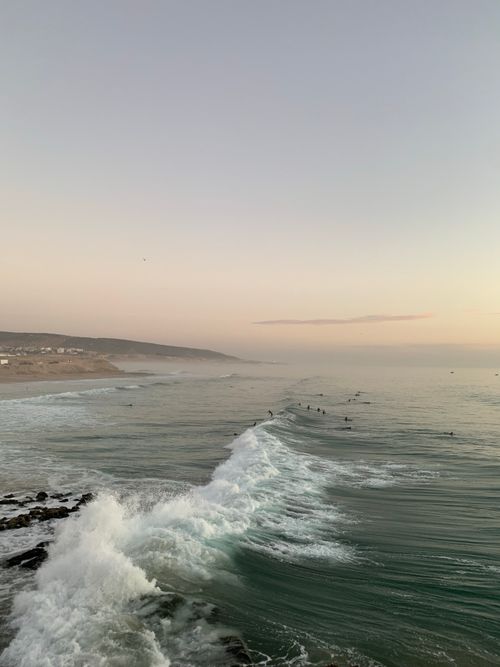 Free Surfers riding waves at sunset on a serene Moroccan coast with calm sea and distant hills. Stock Photo