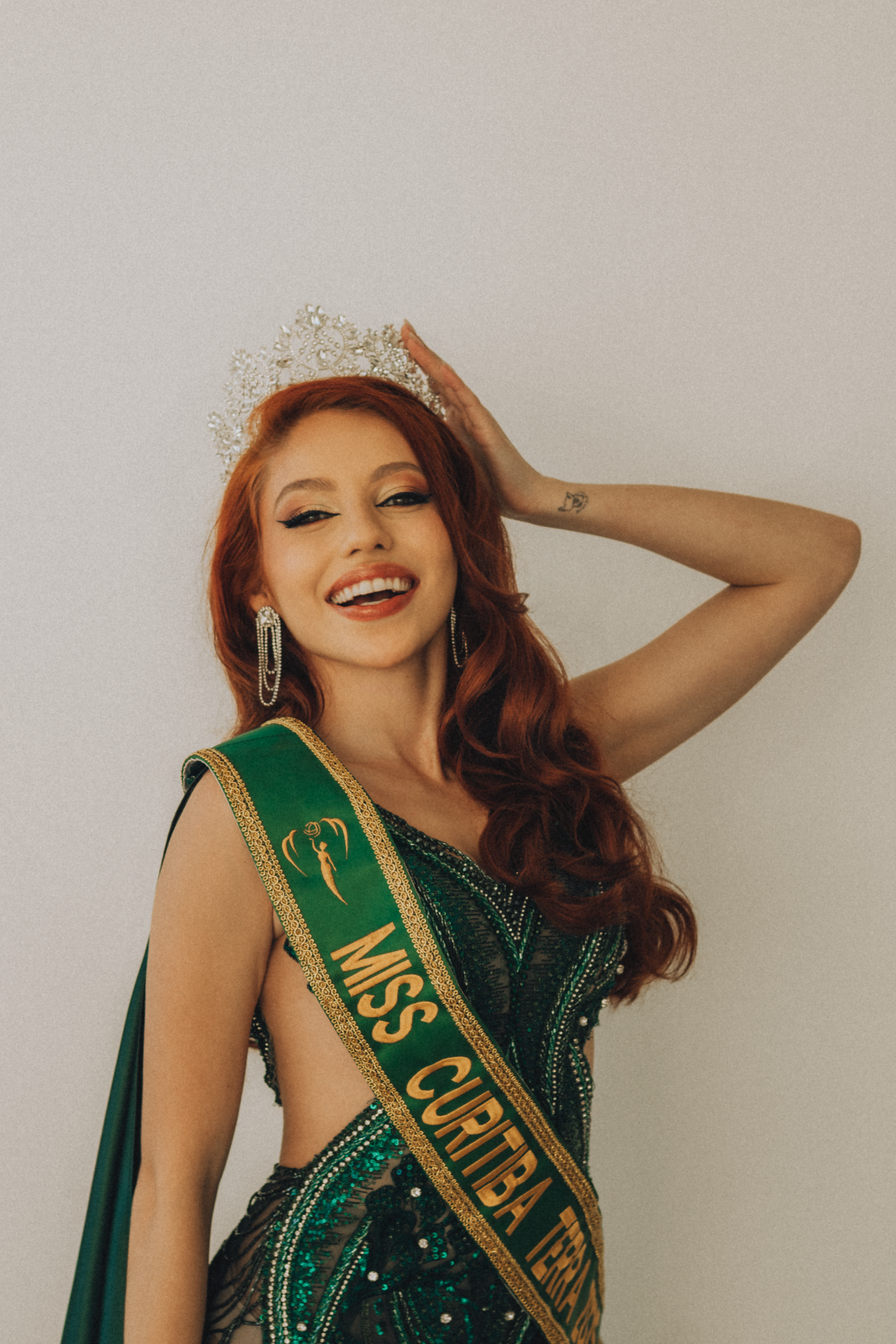 Red-haired woman in a green gown with Miss Curitiba sash, smiling and holding a crown.