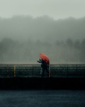A solitary figure walks on a bridge with a red umbrella during misty weather.