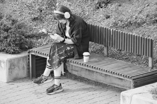 A young woman sitting on a bench listening to music with headphones, enjoying coffee.