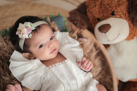 A cute baby wearing a floral headband lies next to a teddy bear, creating a charming and cozy scene.