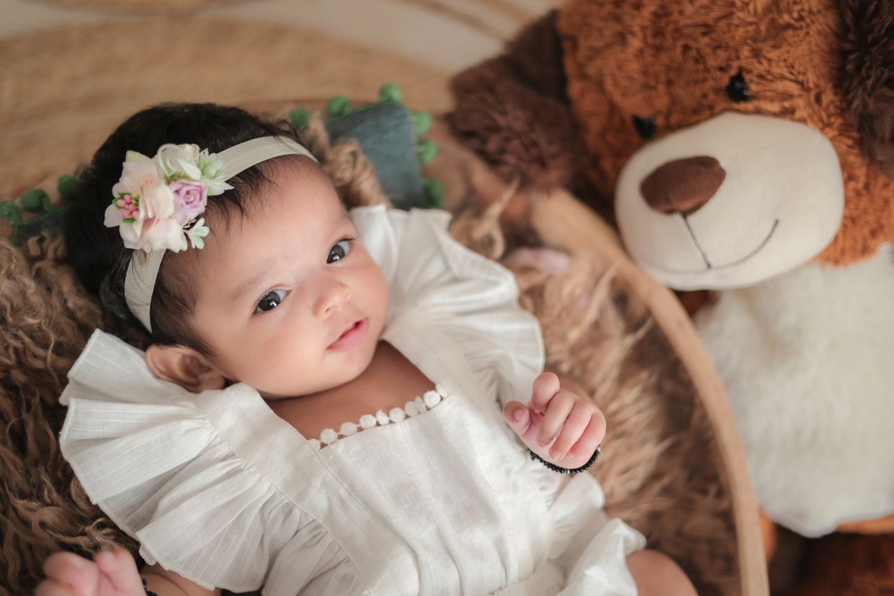 a cute baby wearing a floral headband lies next to a teddy bear creati