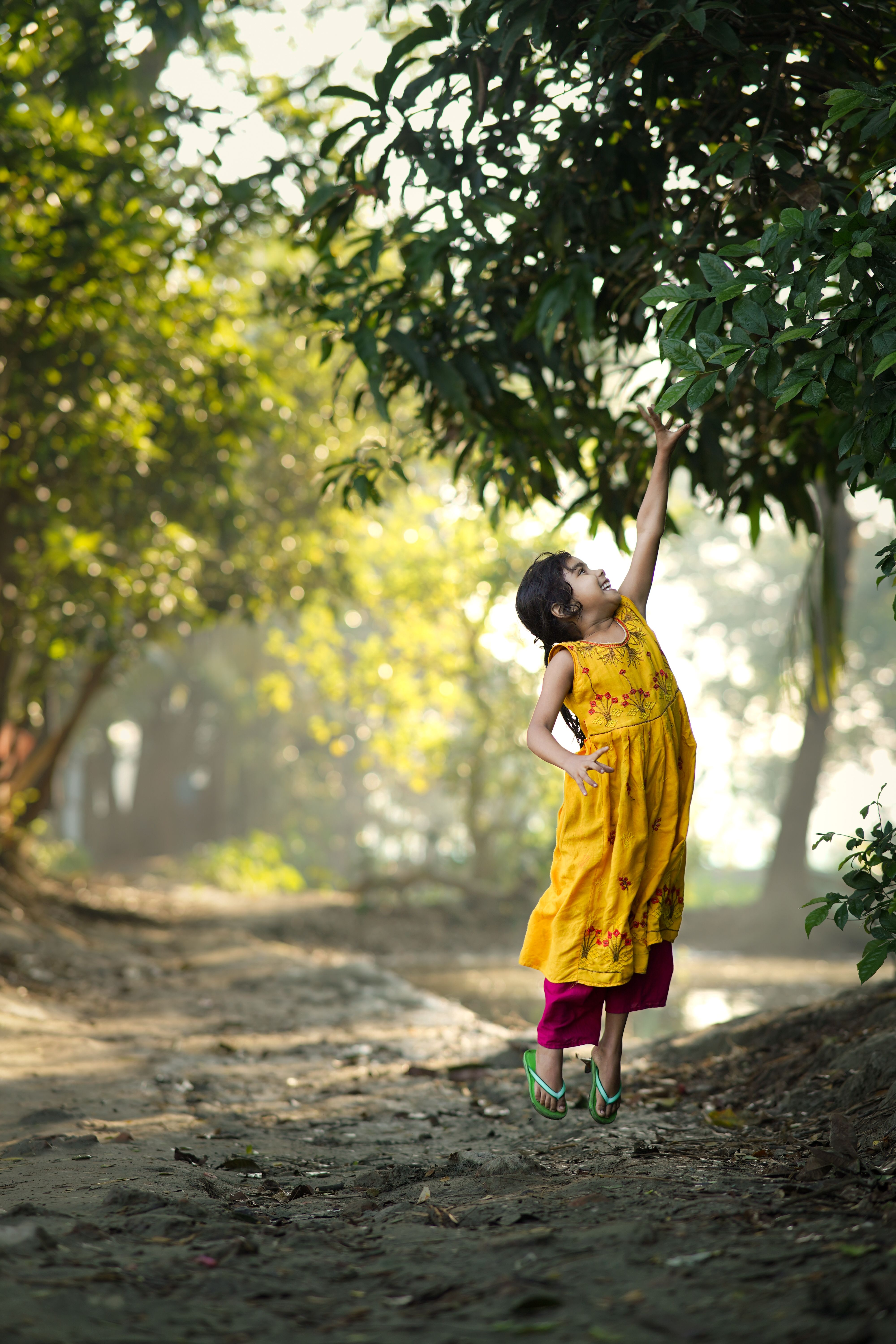 A happy girl reaches for leaves in a sunlit outdoor scene in Bangladesh.