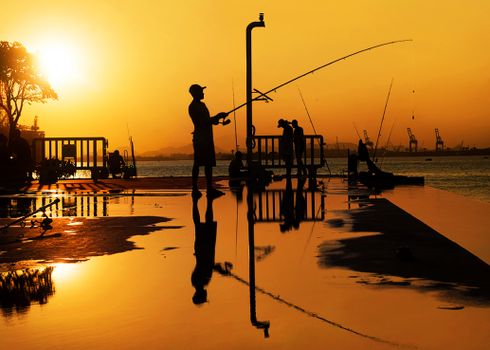 Fishermen cast silhouettes against a glowing orange sunset in Rio de Janeiro, creating a serene and reflective scene.