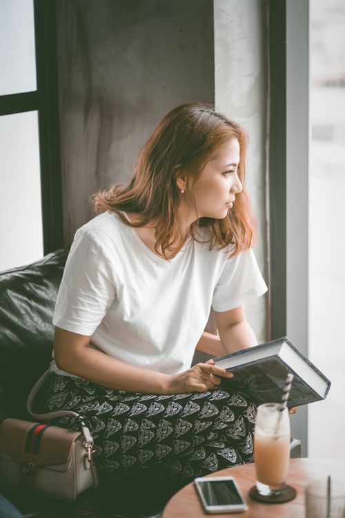 Free A woman sitting by a window reading a book with a smartphone on the table. Stock Photo