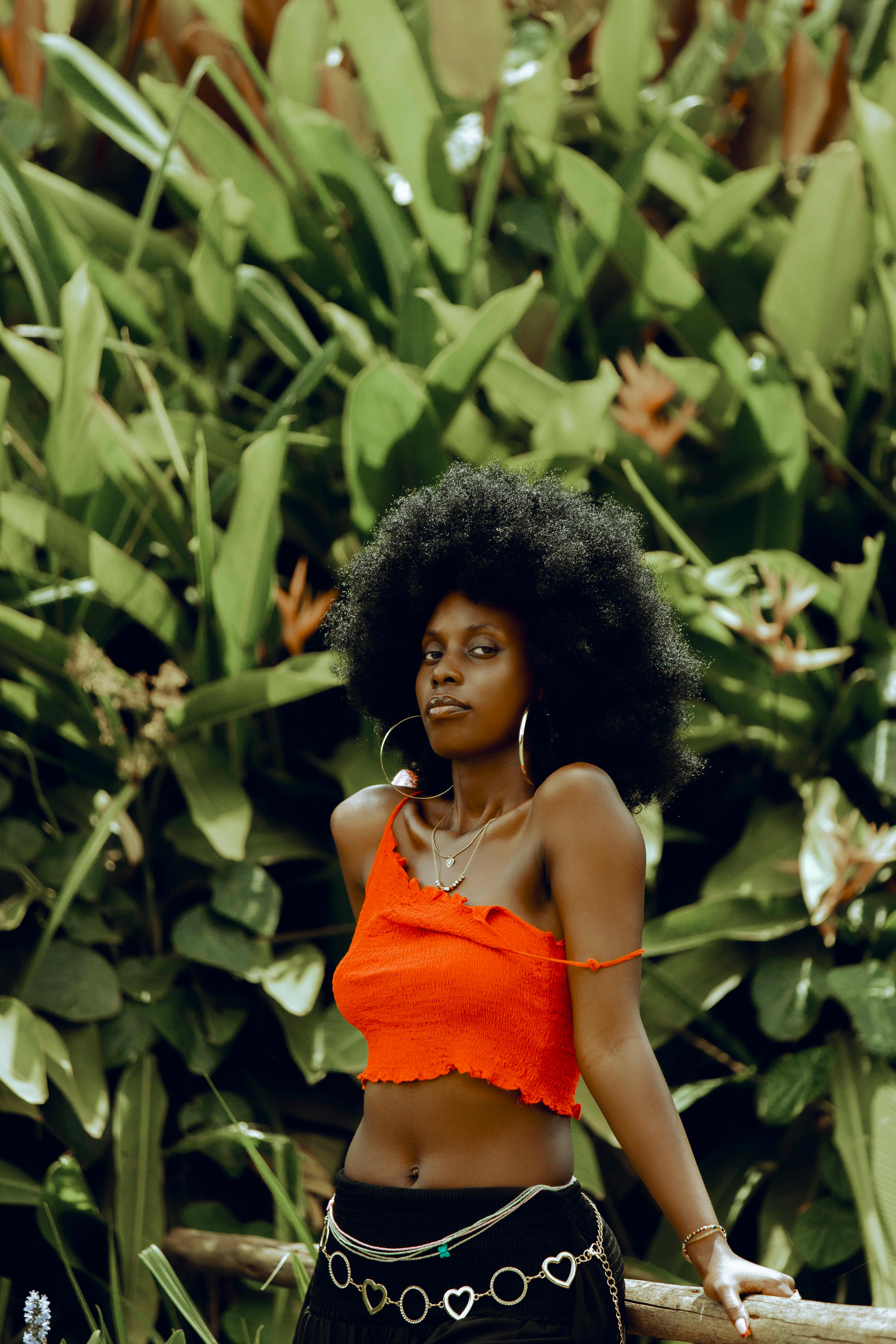 Stylish woman in red top posing amidst lush greenery in outdoor setting.