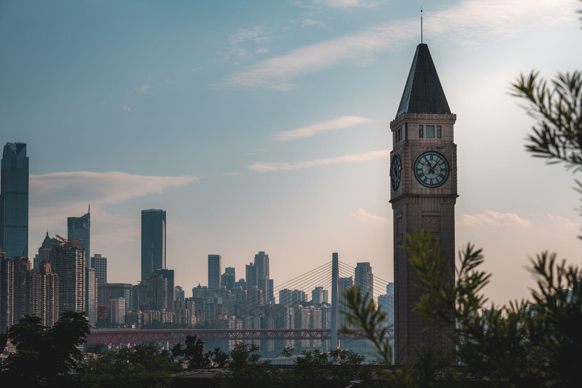 Dramatic city skyline featuring a prominent clock tower at sunset, visible through trees.