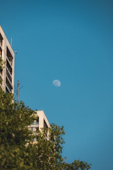 A tranquil daytime urban scene featuring skyscrapers and a visible moon against a clear blue sky.