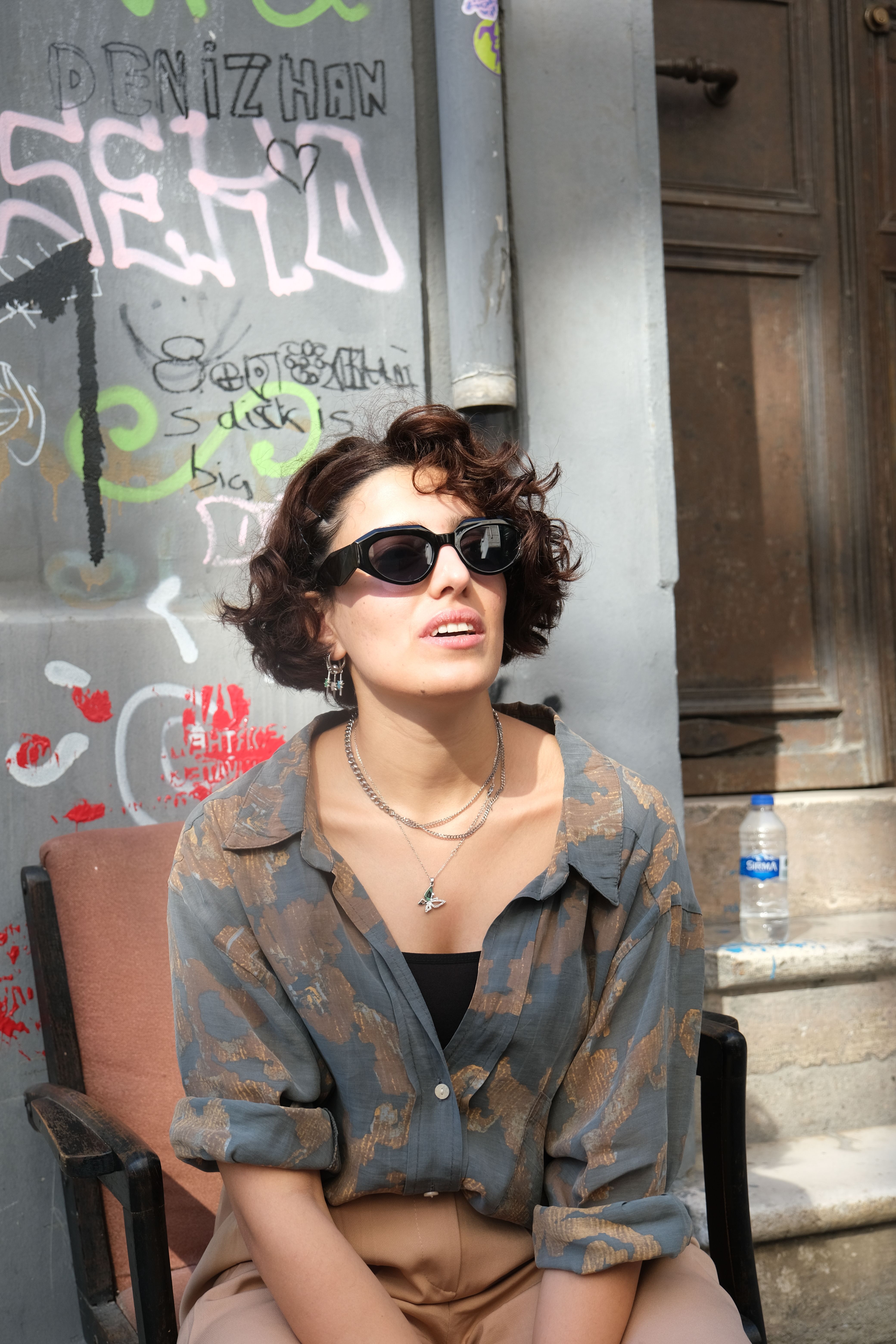 Stylish woman in sunglasses sitting by graffiti-covered wall in urban setting with sunlight accentuating her fashion.