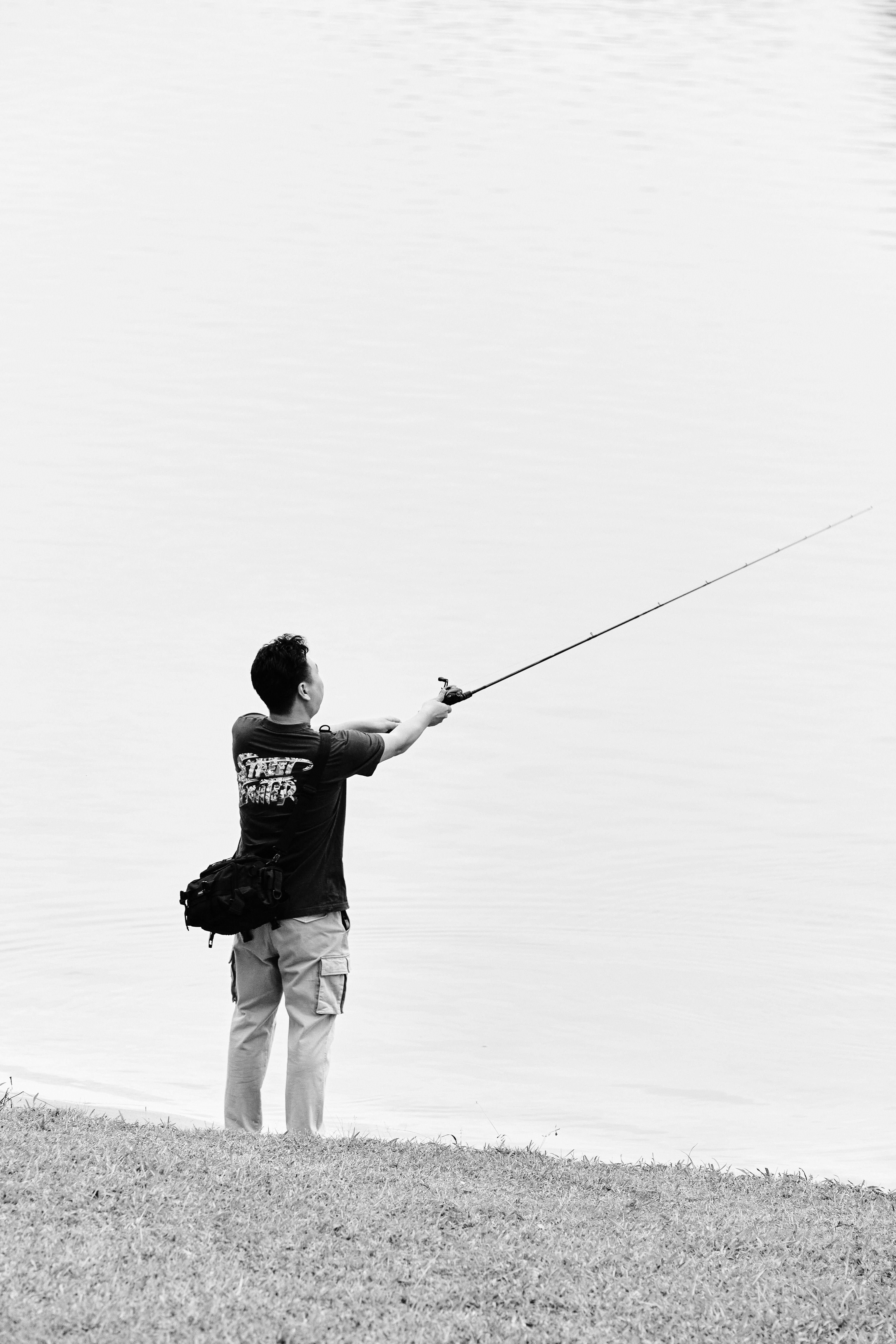 A person fishing by a calm lake in a black and white setting, casting a line into the water.