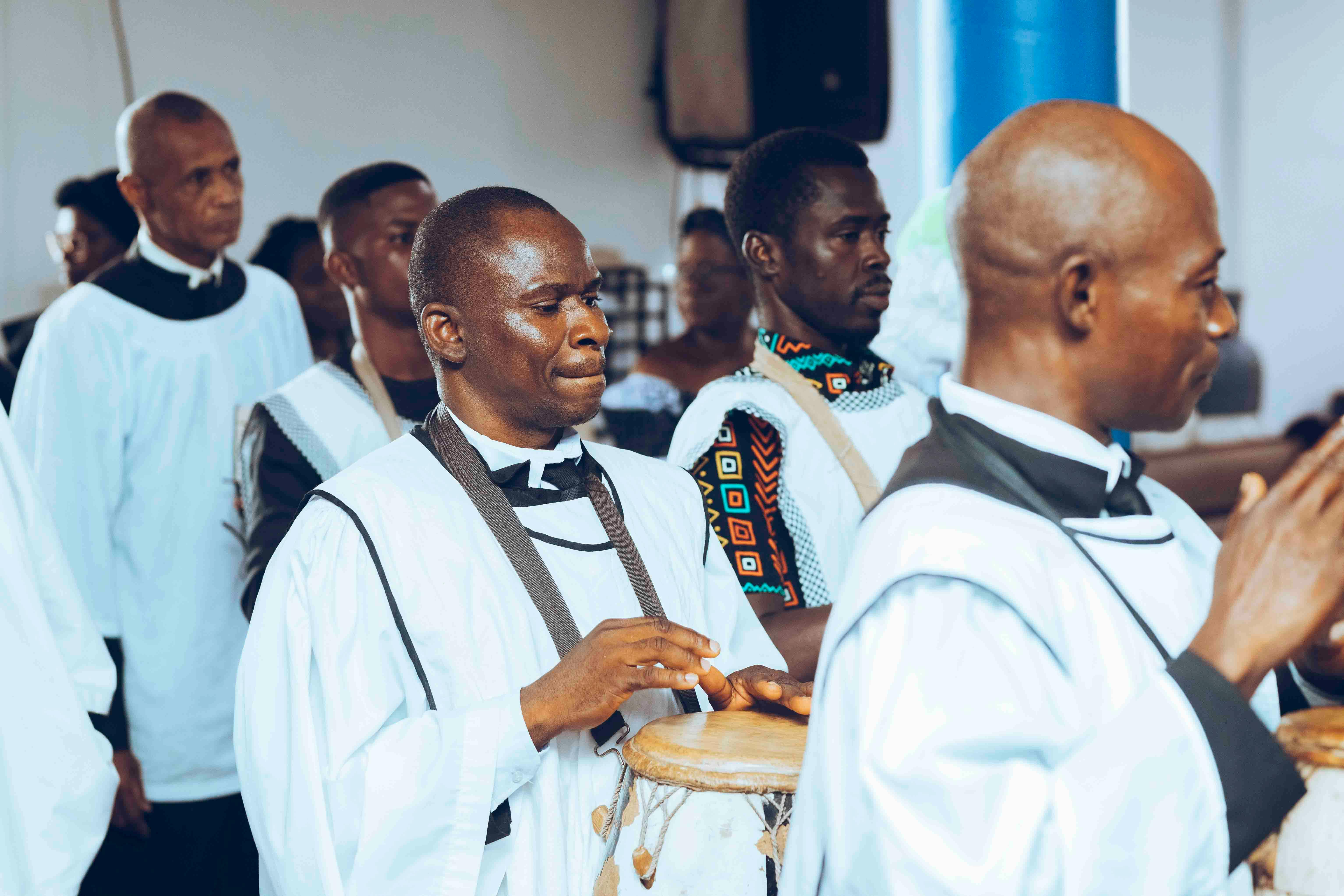 Group of men in traditional African attire participating in a drumming ceremony indoors.