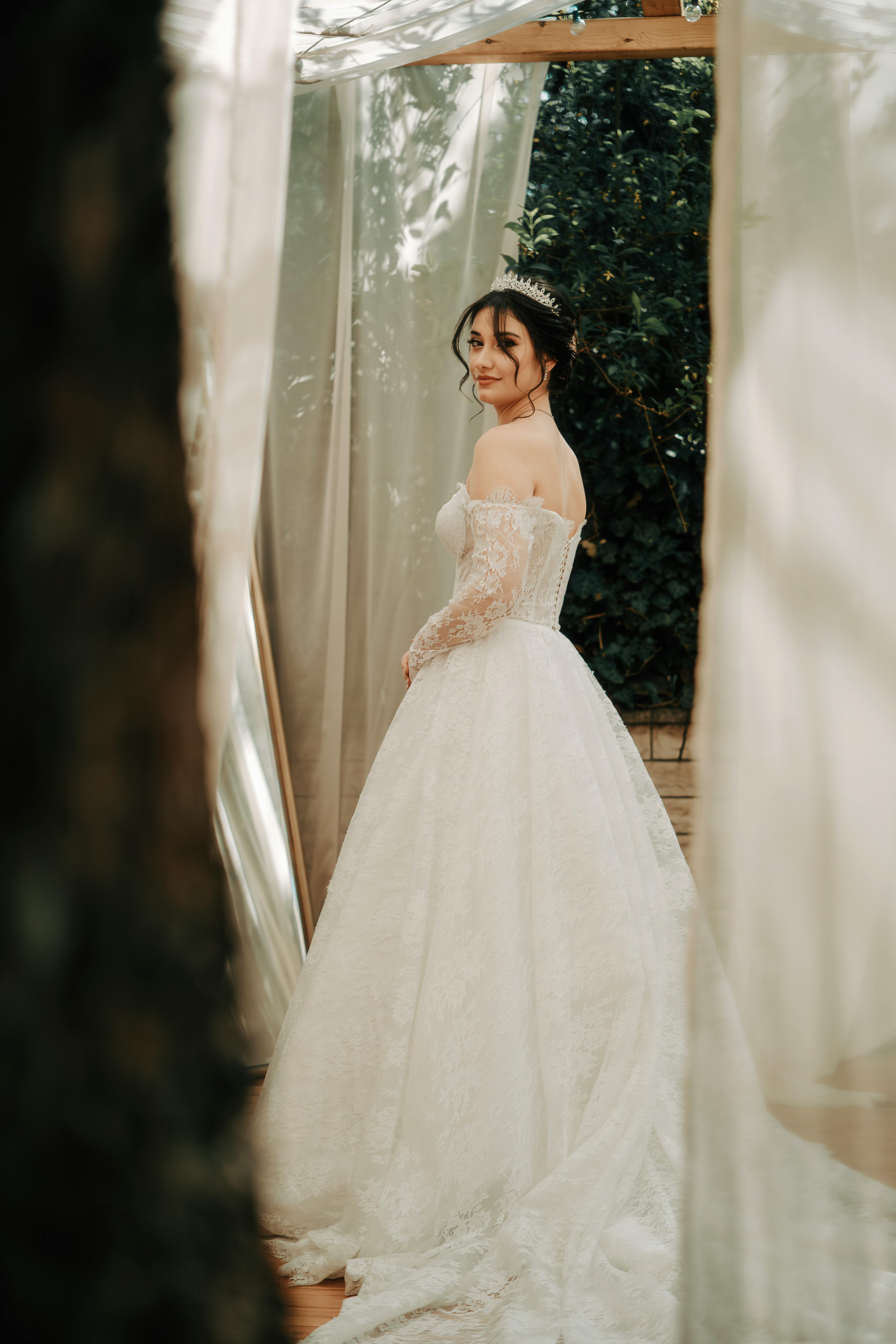 Bride wearing an elegant white lace gown stands outdoors, smiling