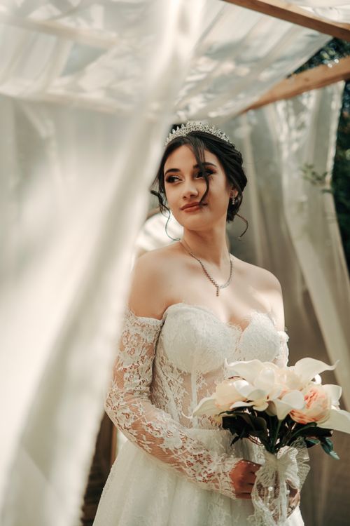 Free A beautiful bride in an elegant lace dress stands gracefully holding a flower bouquet, illuminated by soft sunlight. Stock Photo