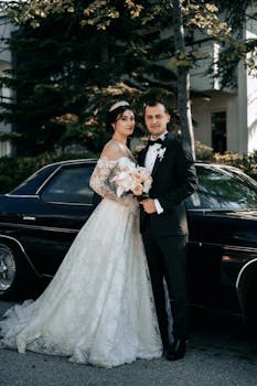 Bride and groom pose elegantly outdoors beside a vintage car.