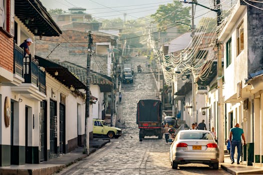 A lively cobblestone street with people, cars, and vibrant buildings during the day in a Latin American town.