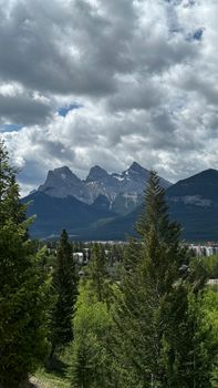 Cloud-covered Three Sisters Mountains in Alberta with lush foreground trees under a dynamic sky.