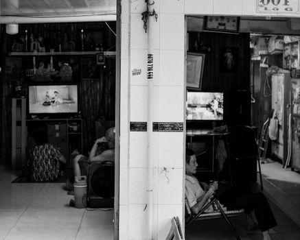 Black and white candid shot of people inside a Vietnamese home, watching TV, depicting daily life.