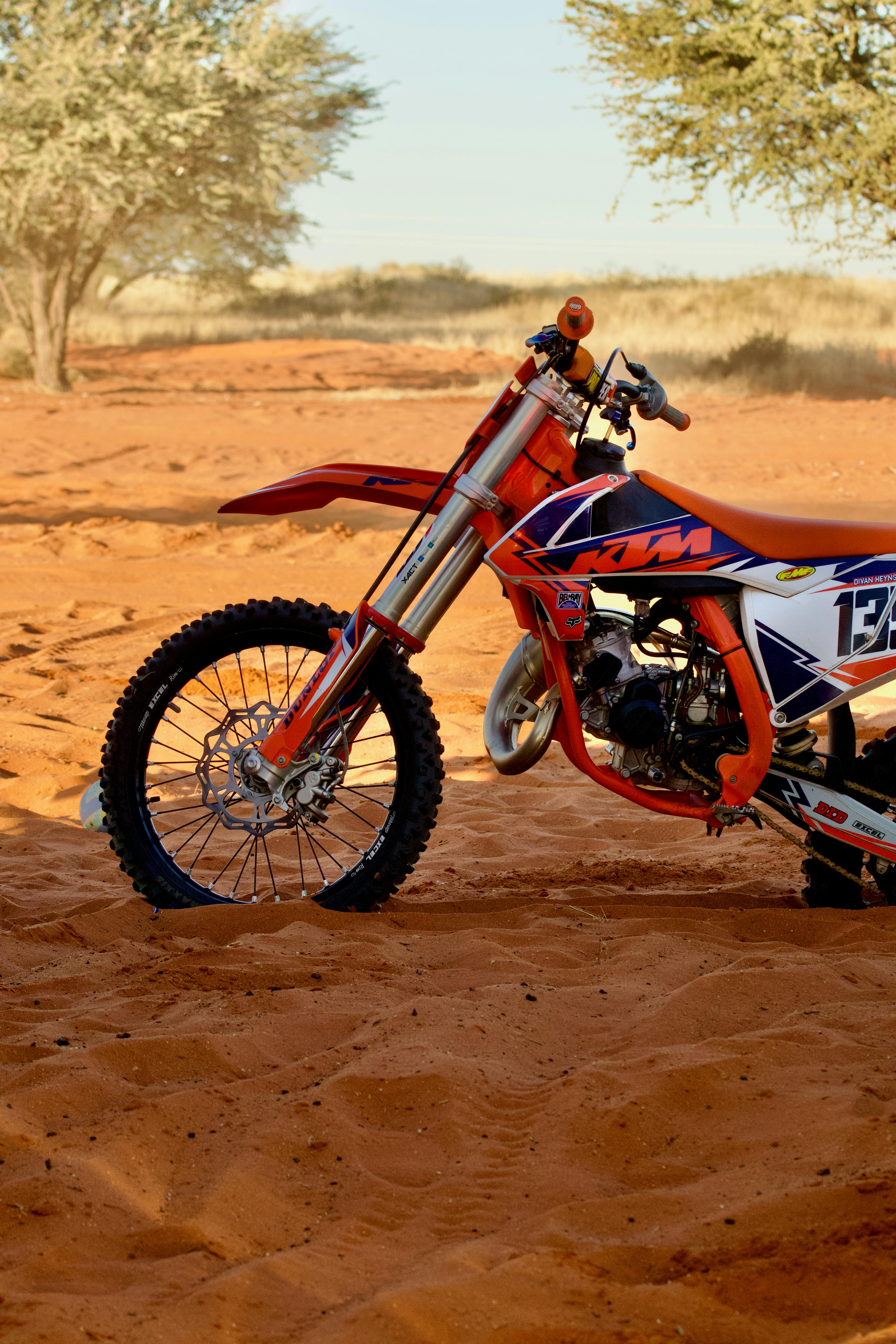 A vibrant dirt bike resting on red desert sand during sunset, surrounded by dry brush and open sky.
