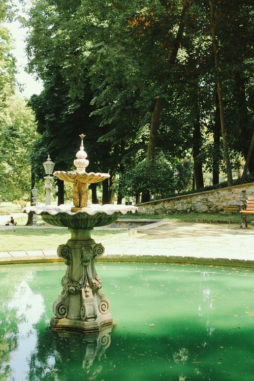 Free A tranquil fountain surrounded by lush greenery in a sunlit park environment, perfect for relaxation. Stock Photo