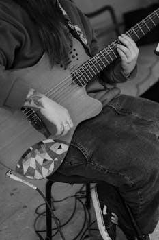 Monochrome image of a person playing an acoustic guitar, focusing on hands and tattoo details.