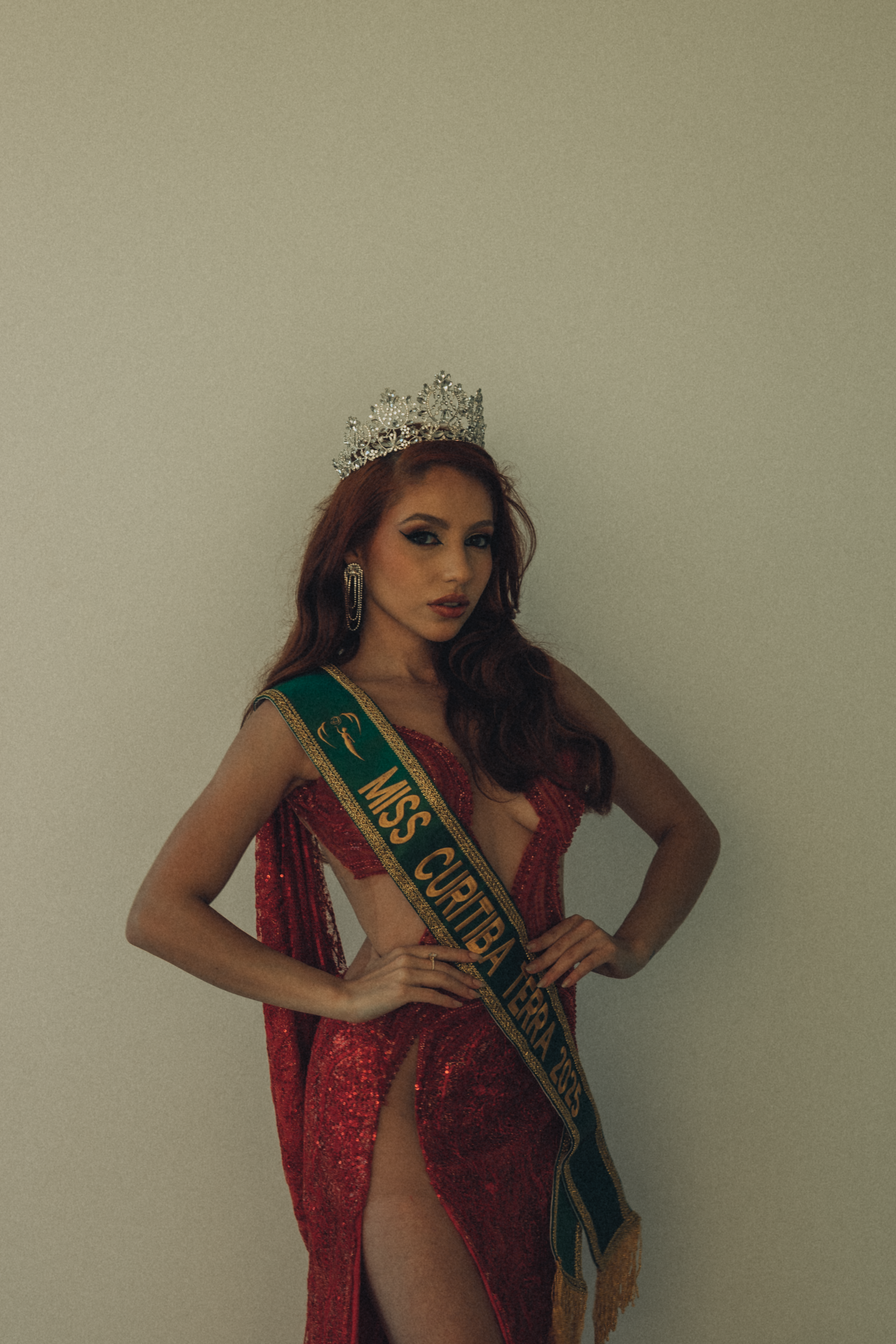 A pageant winner wearing a crown and sash poses in an elegant red dress.