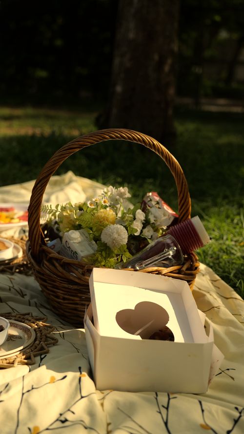 Free Cozy outdoor picnic with a wicker basket, snacks, and flowers on a blanket. Stock Photo
