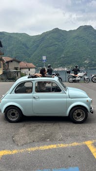 Classic light blue Fiat 500 parked by Lake Como with mountains. Perfect for travel and vintage car lovers.