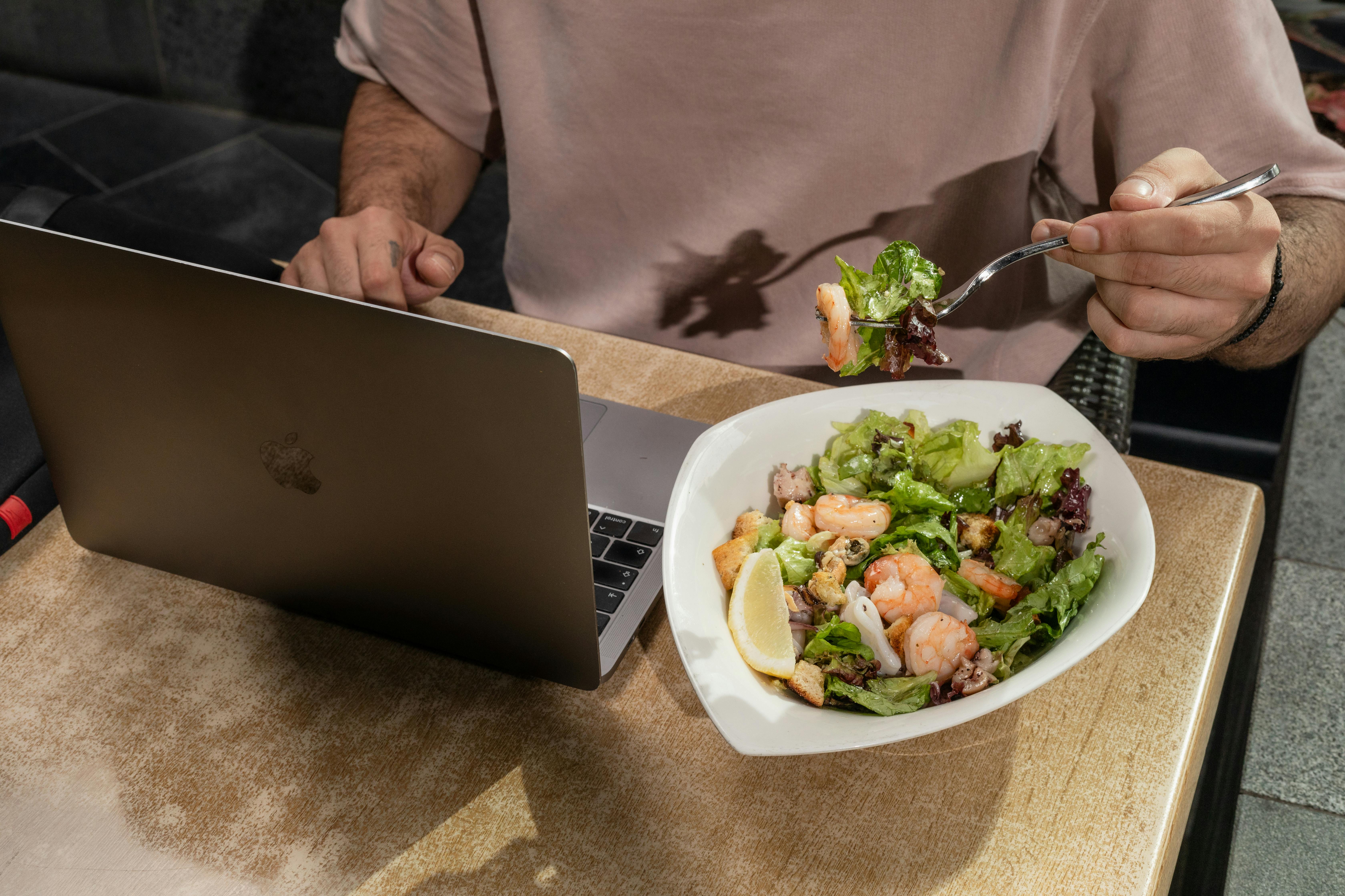 Free Adult man enjoying a shrimp salad while working on a laptop indoors. Stock Photo