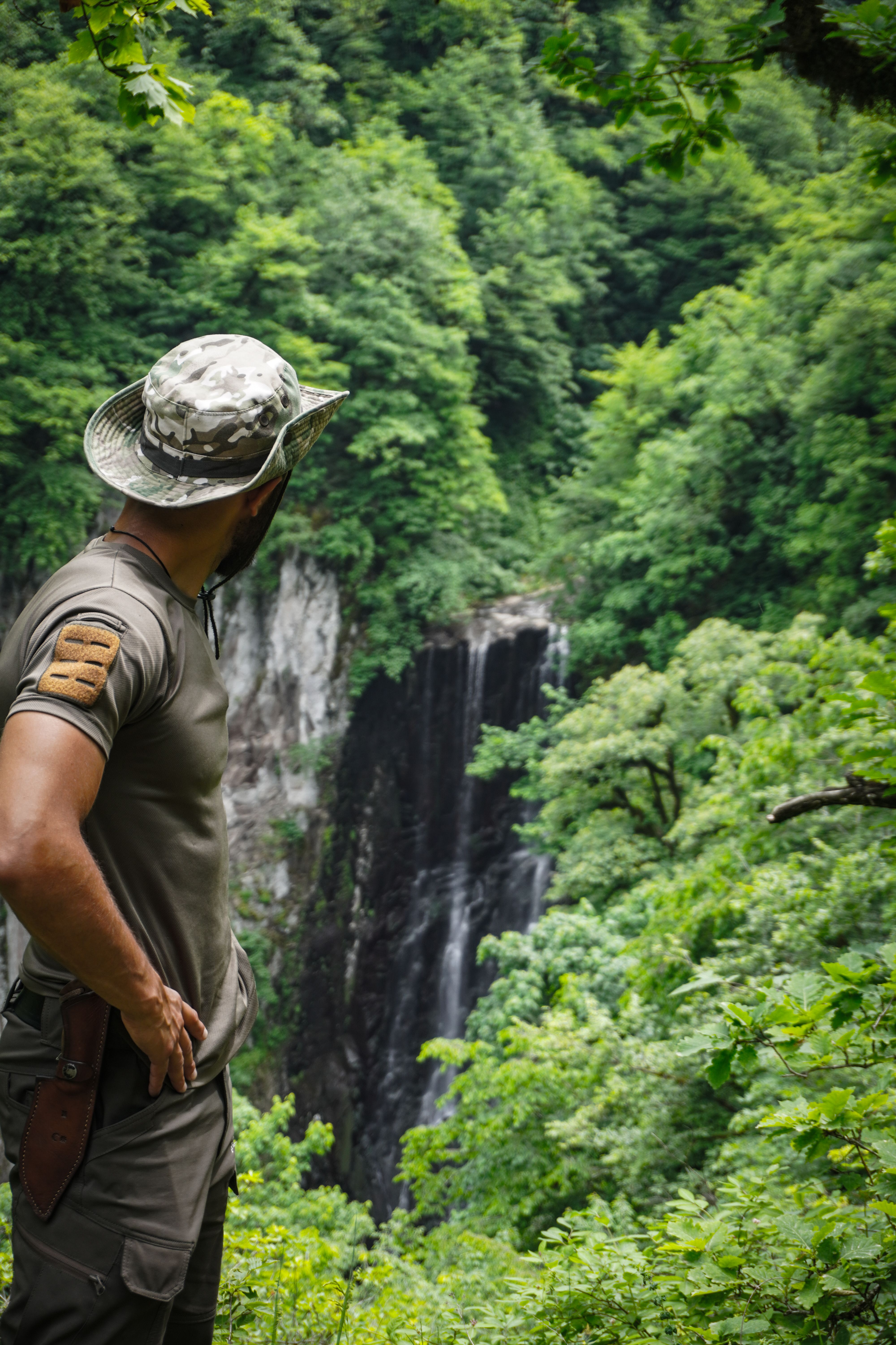 Man in camouflage gear overlooks a stunning waterfall surrounded by dense forest.