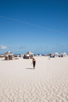 A woman strolls on a sandy beach with beach huts under a clear blue sky. Ideal for travel themes.