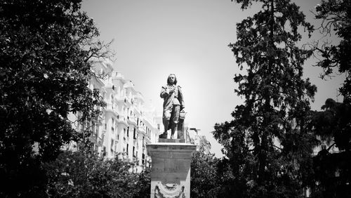 Free Black and white photo of a statue amidst trees and buildings, capturing urban charm. Stock Photo