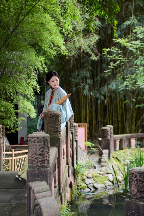 Free A woman in traditional attire stands on a stone bridge amidst lush bamboo forest scenery. Stock Photo