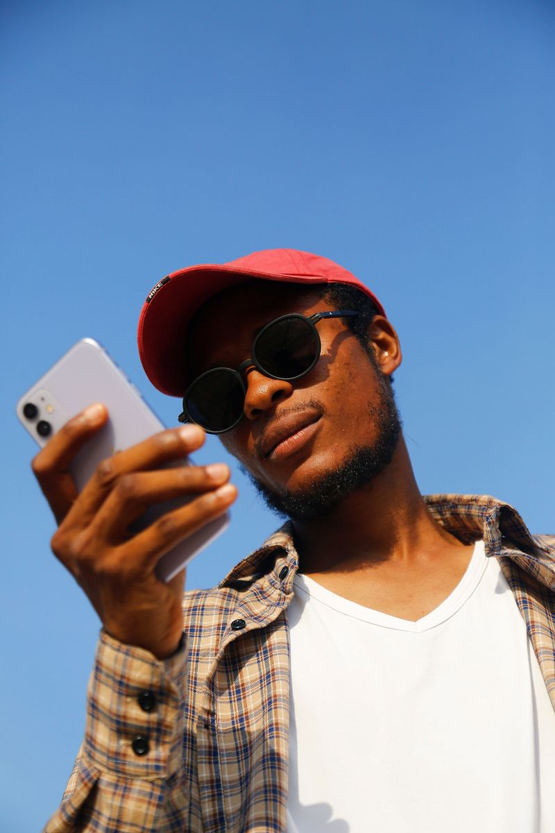Young man wearing sunglasses using a smartphone against a blue sky — photo by Muhammad-Taha Ibrahim on Pexels