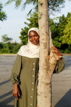 A woman in a hijab smiles behind a tree in an outdoor setting, exuding warmth and confidence.