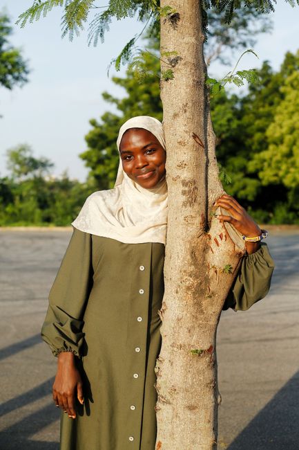 A woman in a hijab smiles behind a tree in an outdoor setting, exuding warmth and confidence.
