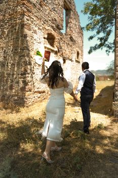 Bride and groom walk near rustic ruins, holding flowers and book passionately.