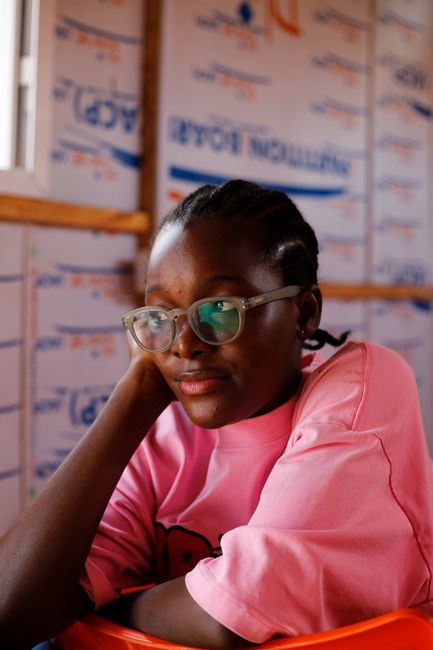 Relaxed young woman sitting indoors, smiling gently, wearing glasses and a pink shirt.