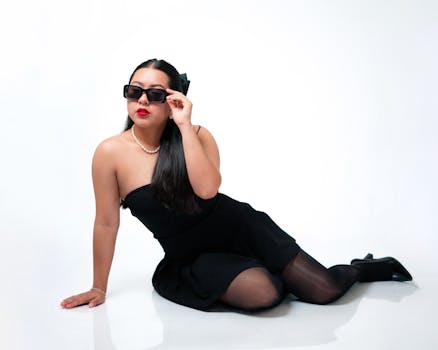 Elegant woman in black dress and sunglasses with a chic pose on white background.