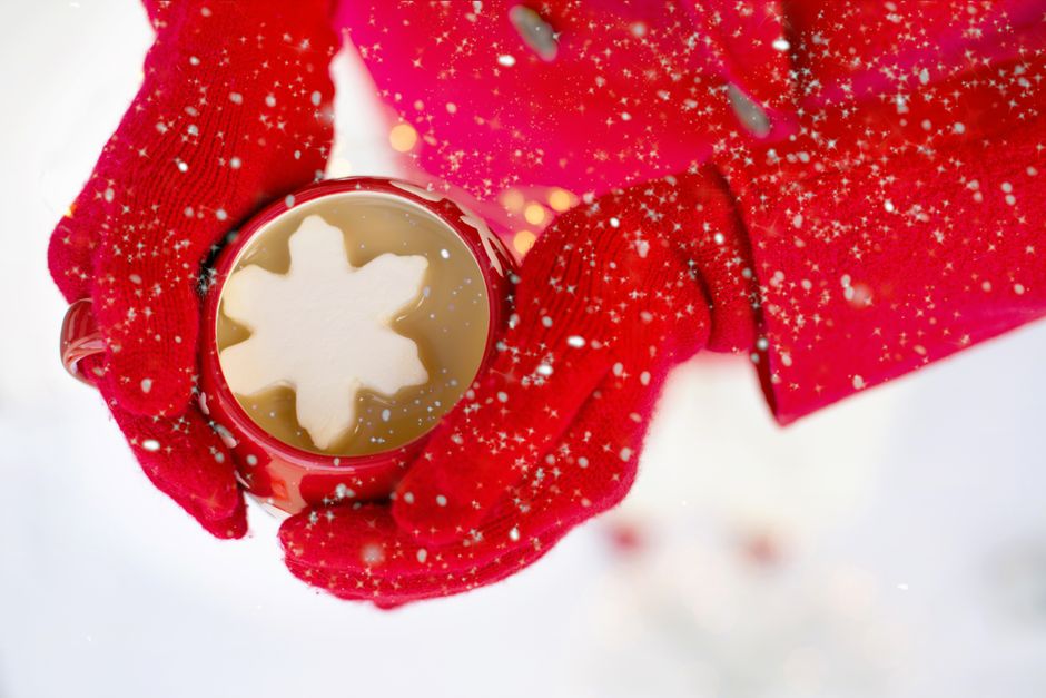 Close-up of red mittens holding a hot drink with a snowflake marshmallow in winter snow.