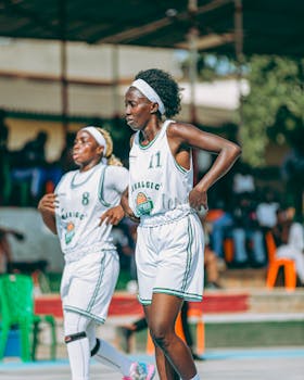 Two women basketball players in a match outdoors wearing white uniforms.
