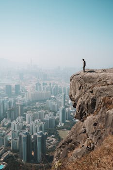 Man stands on cliff overlooking stunning Hong Kong skyline, capturing the blend of urban and natural environments.