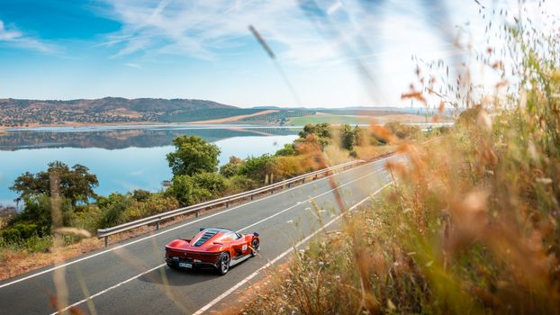 A stunning red sports car cruising on a picturesque road beside a serene lake under a clear blue sky.