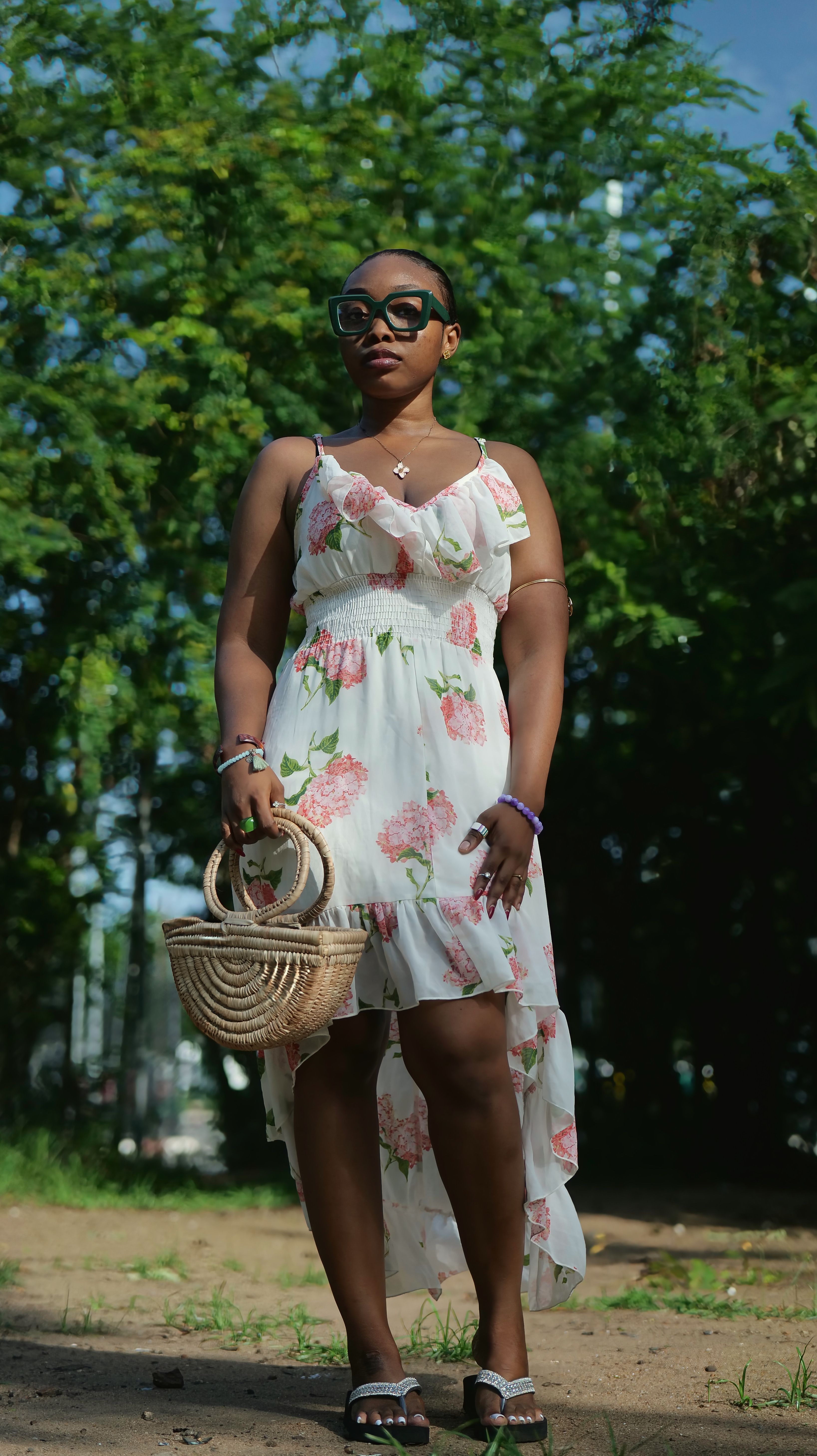 Fashionable woman in floral dress and sunglasses stands outdoors carrying a woven bag.