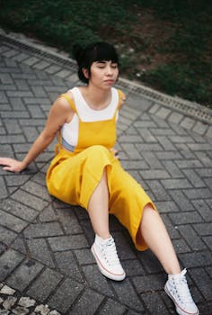Woman in yellow dress sits on a paved pathway outdoors, wearing white sneakers.