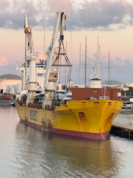 Yellow cargo ship docked at harbor during sunrise with industrial cranes and calm water.