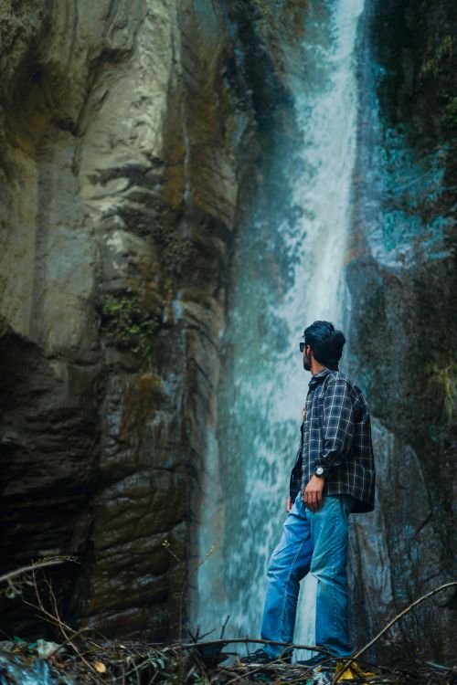Free A man stands at the base of a waterfall in nature, surrounded by rocky cliffs. Stock Photo